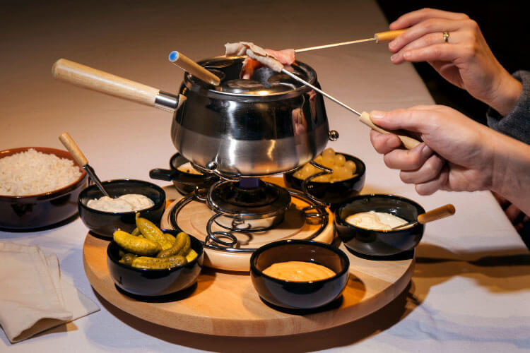 Two hands cook pieces of meat in a fondue pot. Six dishes of dipping sauces surround the pot of Fondue Chinoise.