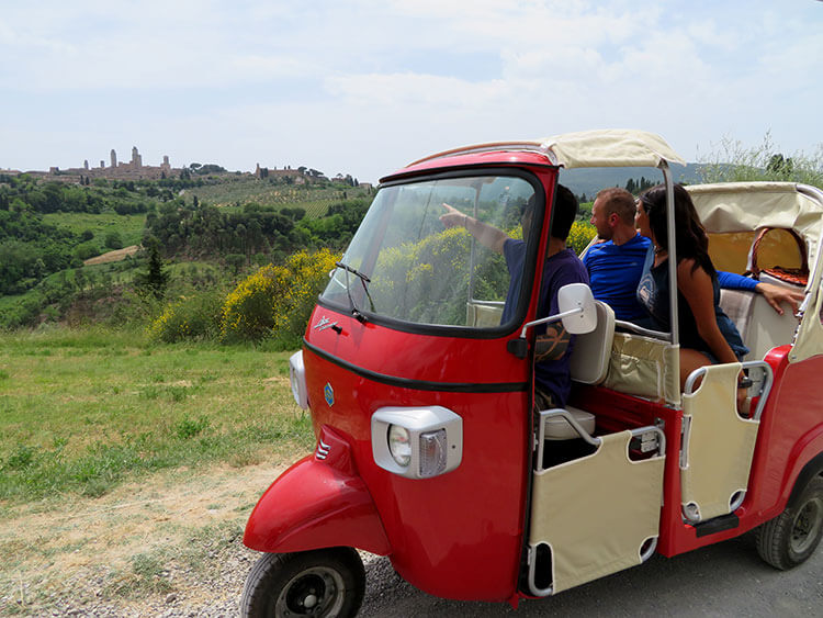 The guide points toward San Gimignano from a roadside viewpoint of the medieval town during the tuk tuk tour of Tuscany