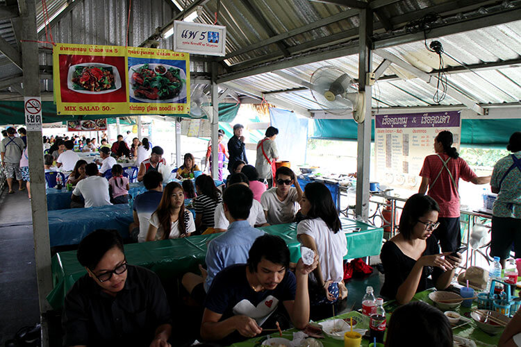 Low tables filled with Thai locals eating various foods and gathering socially at the Taling Chan Floating Market