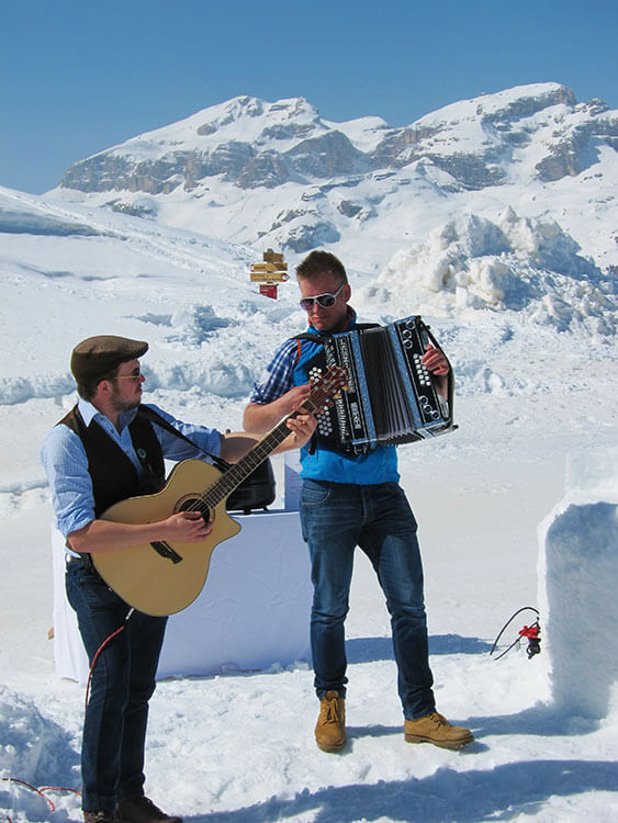 One man plays a guitar while another man plays an accordian with the snowy mountains behind