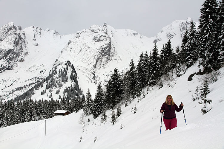 Jennifer snowshoeing with pine trees and the mountains of Massif des Aravis behind