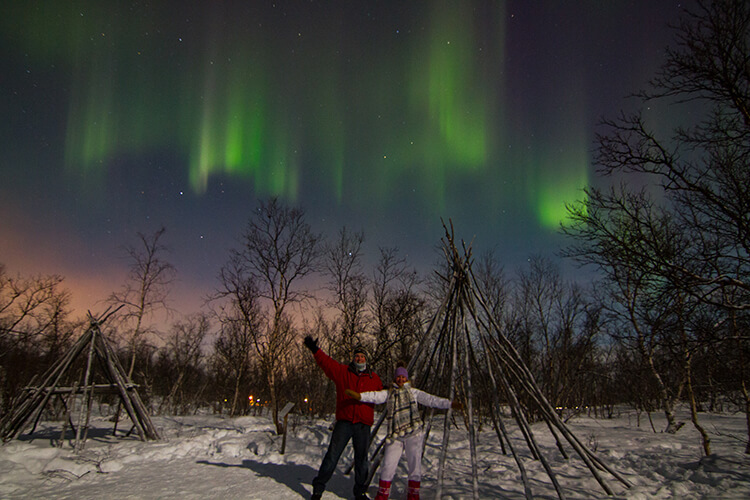 Jennifer and Tim pose under a green and purple curtain of Northern Lights in Abisko, Sweden