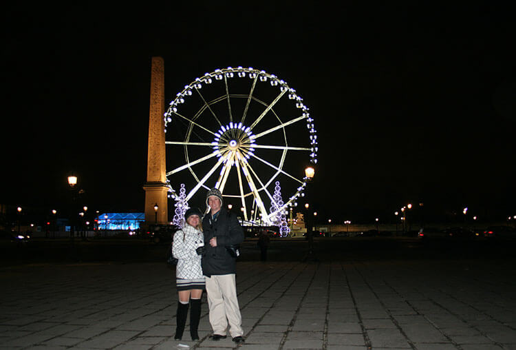 Jen & Tim pose with the Grand Roue a Paris lit up on Place de la Concorde in Paris at Christmas