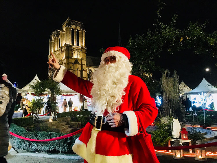 A Santa Claus poses at the Marche de Noel Paris Notre Dame while the cathedral is illuminated behind him