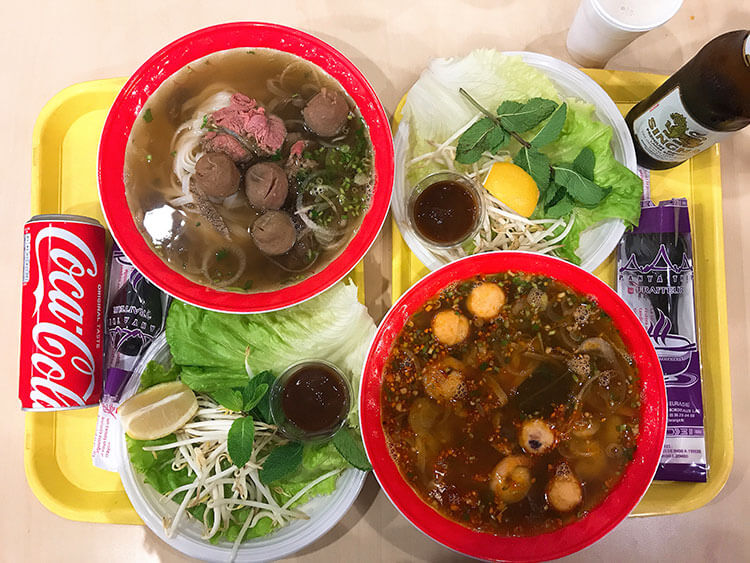 A bowl of pho and a bowl of Tom Yum Goong with plates of herbs and sprouts at Panya Thip Eurasie