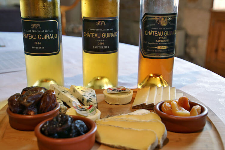 A cheese board with three cheeses, dates, apricots and prunes and bottles of the Chateau Guiraud 2014, 2006 and 1998 vintages