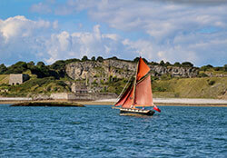 A red sailboat sails past ruins on the Anglesey coast near Beaumaris