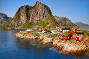Red and green wooden houses of the village of Hamnøy sit on rocks under a vertical mountain in the Lofoten Islands