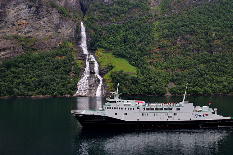 The multi-tiered The Suitor waterfall tumbles down the rocky cliffs of the fjord and a ferry passes in front in Geiranger
