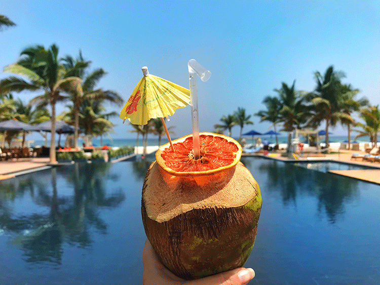 Jennifer holding up a fresh coconut at the main infinity pool at Al Baleed Resort by Anantara