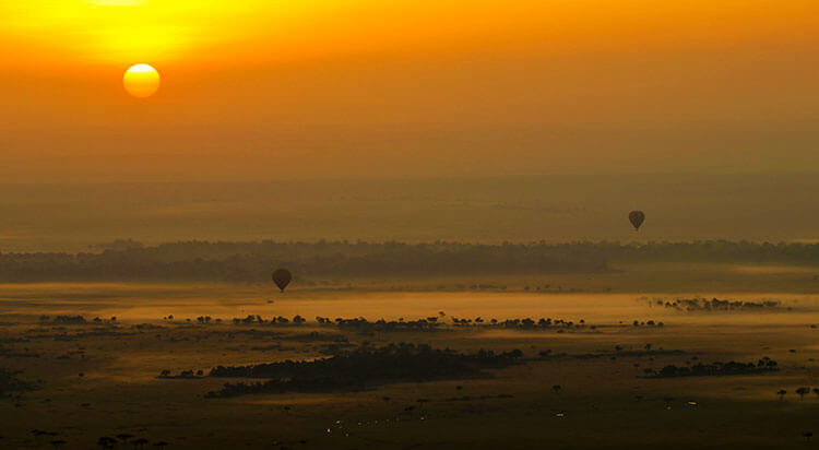 Two hot air balloons float above the Mara River as the sky turns shades of orange and the sun appears as a fiery ball at sunrise