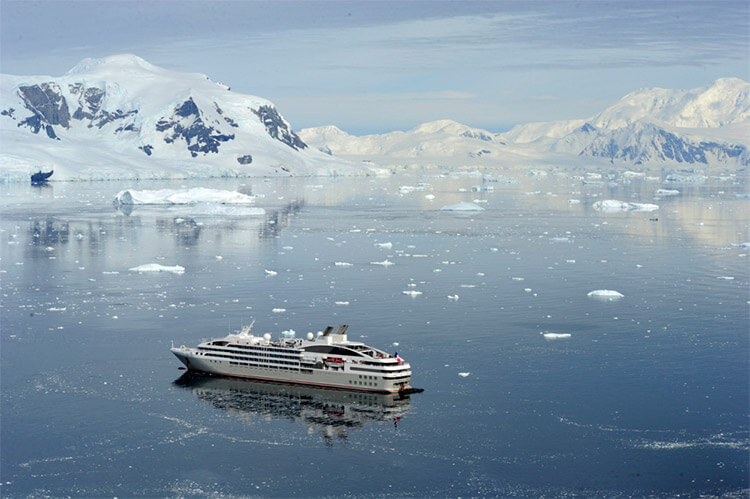 A PONANT ship surrounded by the icebergs with mountains rising up along the shores in the distance in Antarctica
