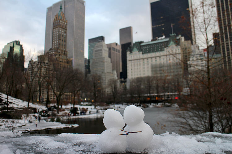 Two tiny snowmen on the Gapstow Bridge with sticks to make it look like they are hugging with Central Park South and the Plaza Hotel as the backdrop