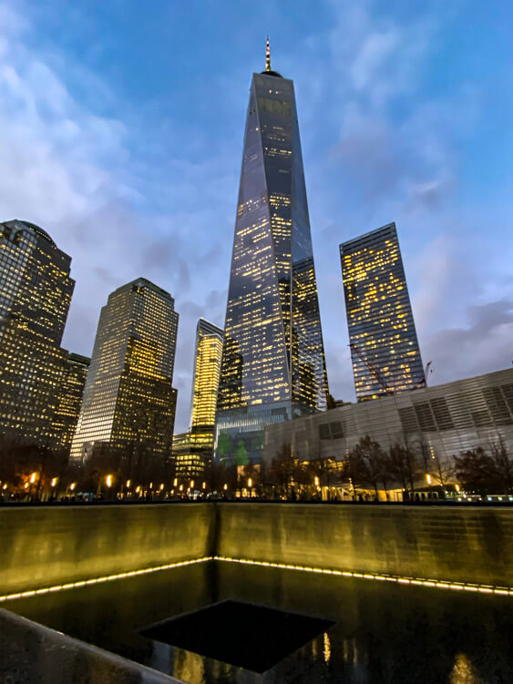 One World Trade Center stands lit up at dusk behind one of the reflecting pools of the 9/11 Memorial 