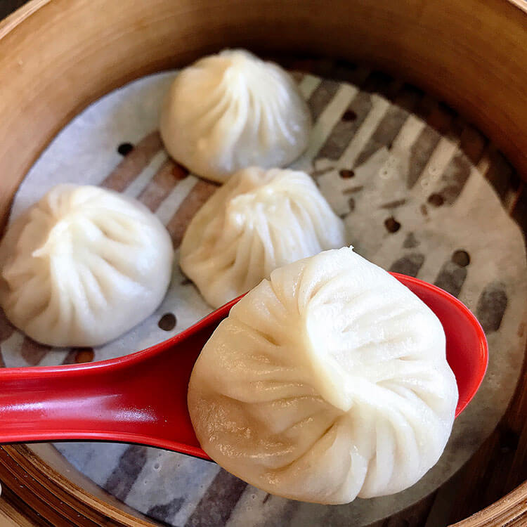 Holding a soup dumpling on the spoon over the steamer basket with more soup dumplings in it