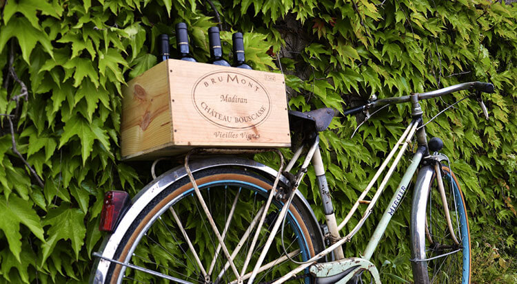 A bike leans up against bushes and has a basket made from a Vignobles Brumont wine box and is filled with wine bottles