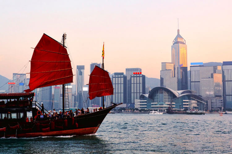 A red sail junk boat sails past the modern Hong Kong skyline