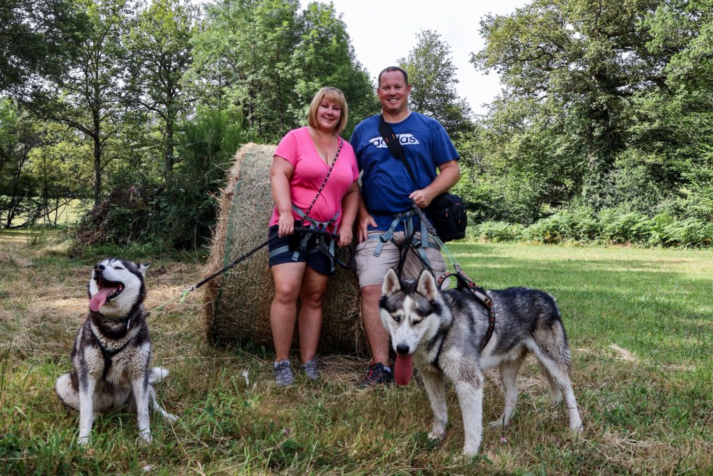 Jennifer and Tim lean against a hay bale as the two huskies take a break during a hike with Husk'in Creuse