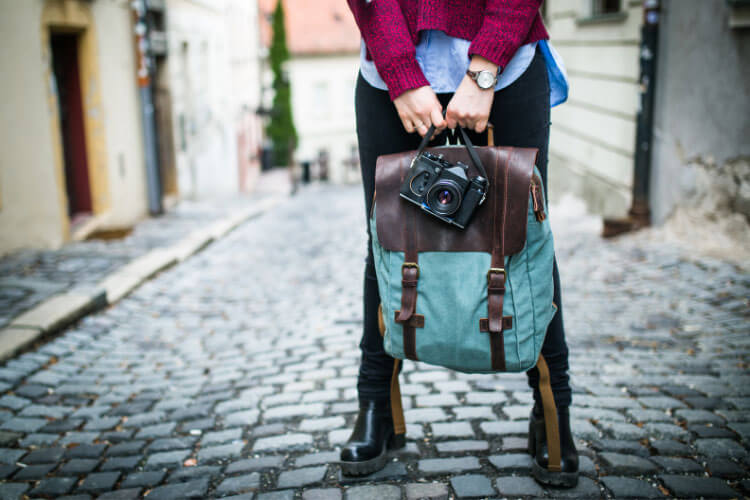 A woman holds a compact camera along with a backpack while standing on a cobbled street