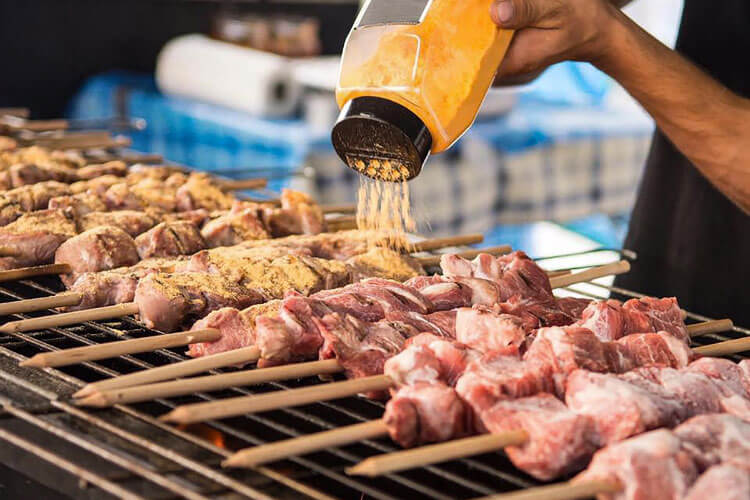 A cook sprinkles seasoning on meats on sticks he's grilling at a cart in Bangkok's Chinatown