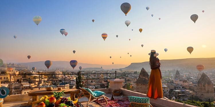 A woman stands on the sun terrace at sunrise watching hot air balloons fill the sky from Mithra Cave Hotel in Cappadocia