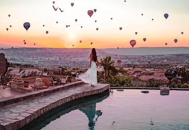 A woman stands at the edge of the pool at Museum Hotel in Uchisar watching sunrise and hundreds of hot air balloons in Cappadocia