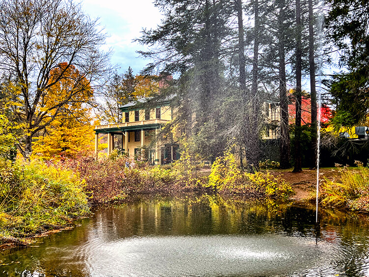 The yellow and green Glen Iris Inn seen tucked into the trees from across the small pond with fountain in Letchworth State Park