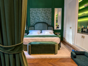 The bedroom of a junior suite at Hotel La Gemma with emerald green walls, botanical palm leaf wallpaper, bedding and curtains in rich emerald velvet