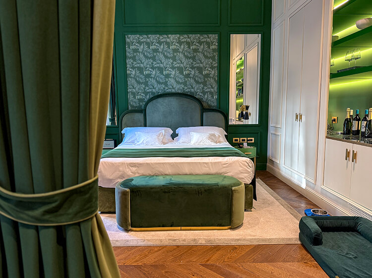 The bedroom of a junior suite at Hotel La Gemma with emerald green walls, botanical palm leaf wallpaper, bedding and curtains in rich emerald velvet