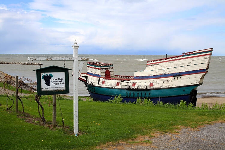 A sign noting the Lake Erie Concord Grape Belt with a beached wooden boat behind at Barcelona Harbor Beach